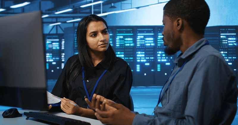 Two IT professionals, a woman and a man, discussing work in front of a computer in a server room.